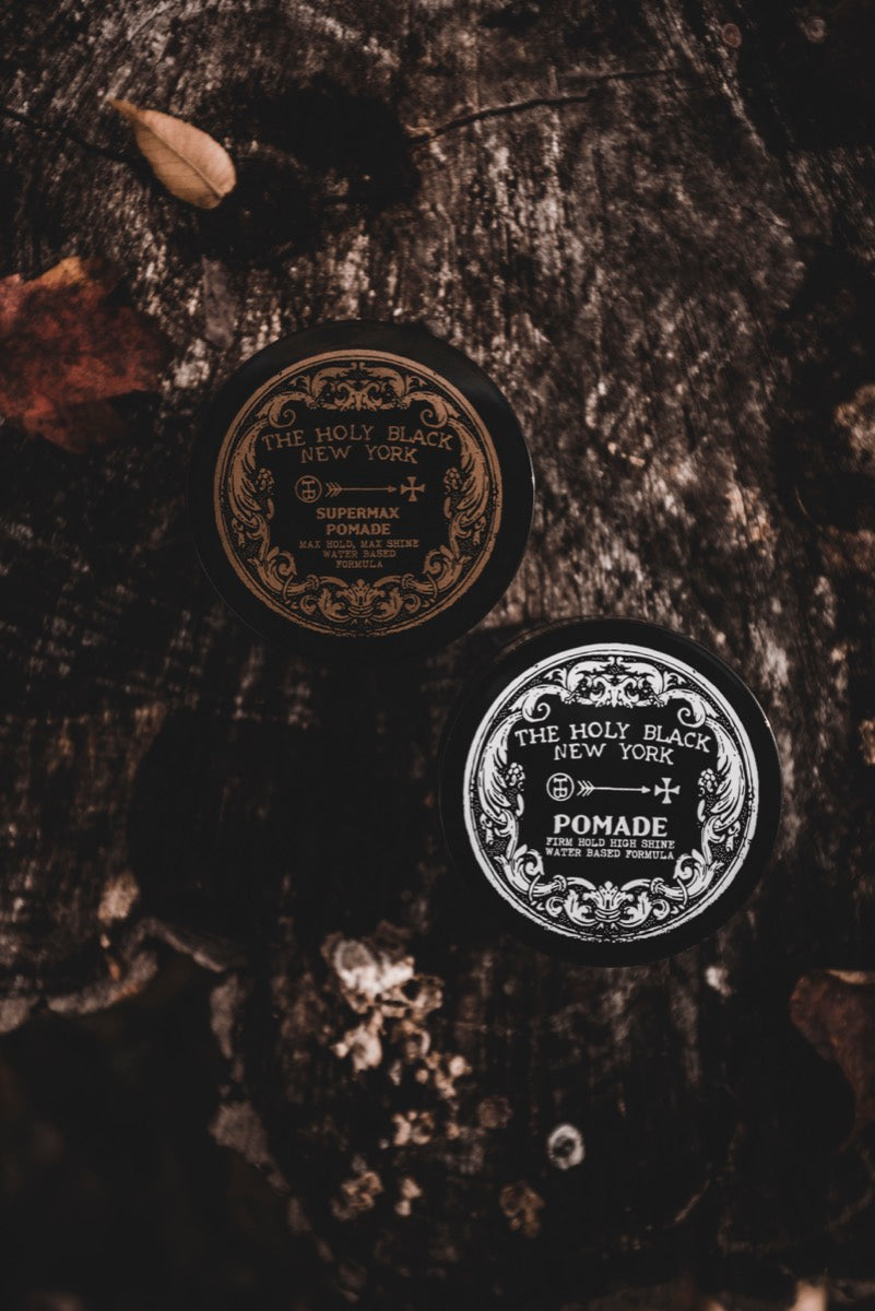 Two Holy Black pomade jars on dark textured surface
