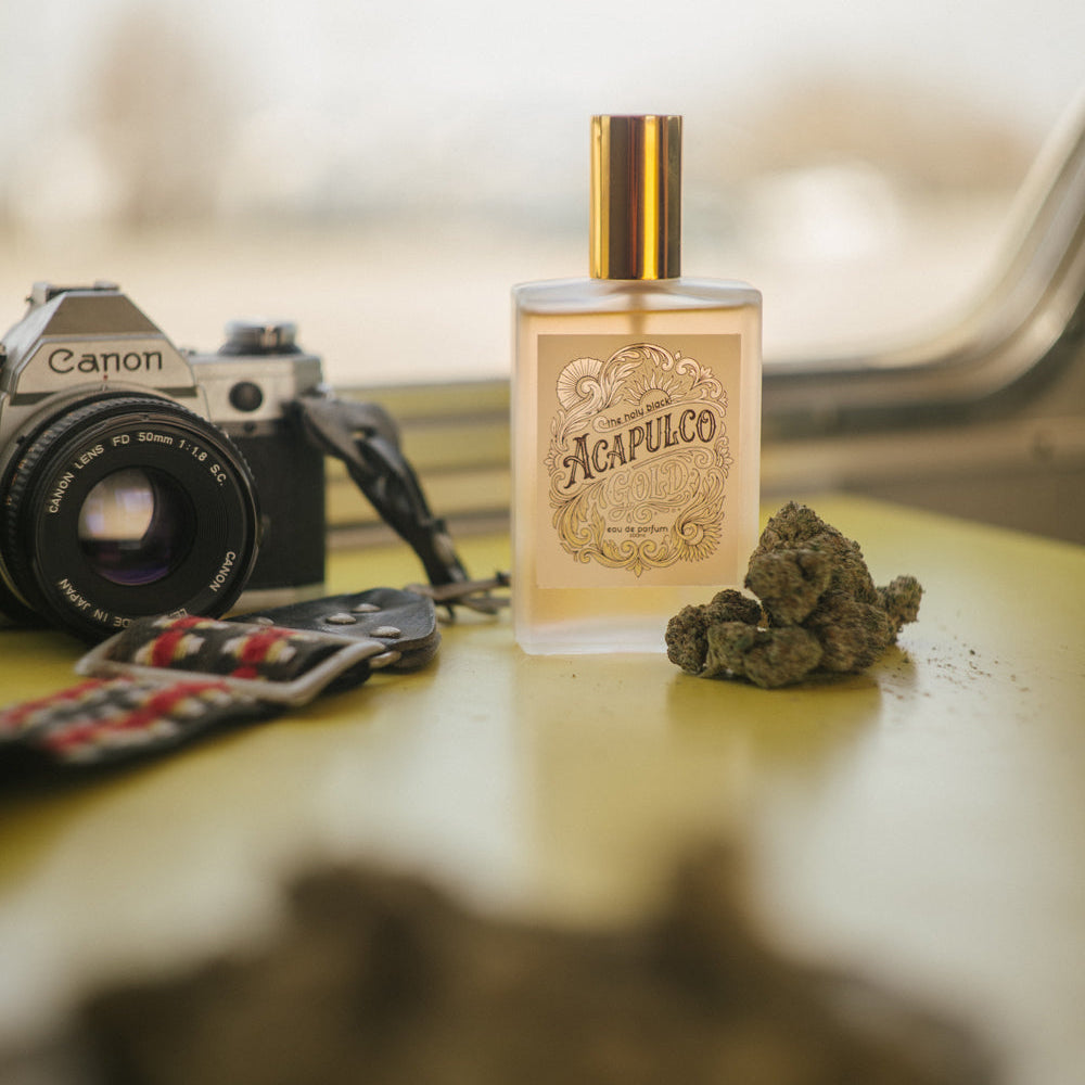 Perfume bottle labeled 'Acapulco' with cannabis buds on a table next to a camera.